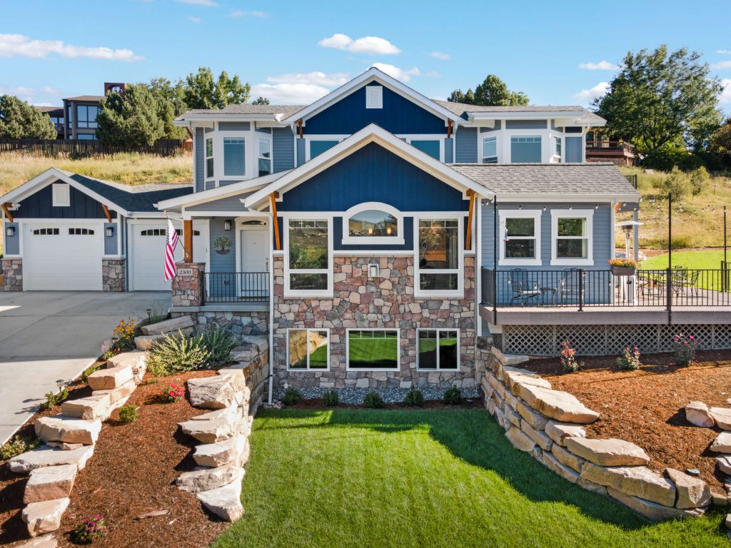 a blue mountain craftsman with large stones framing the yard looks stunning against a rolling green hill. Lots of layers and gables and porches make this one of the stand out new homes in windsor co.